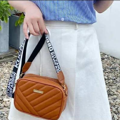 Person holding a brown quilted handbag with a black strap featuring white text.